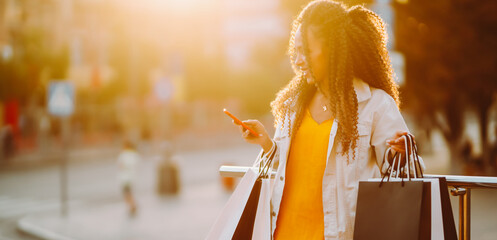 A smiling woman in stylish clothes with shopping bags enjoys the sunny day with stores in the...