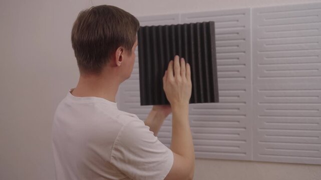 Back view of young adult man comparing different soundproofing acoustic panels and placing them on wall for noise reduction, improving room acoustics for sound recording or home studio, slow motion.