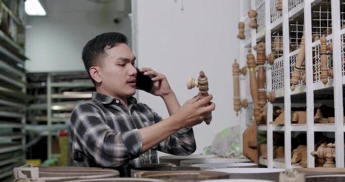Asian adult male woodworking business owner holding wooden spindle while talking on phone, slowly turning material to confirm order details, inside interior construction material shop