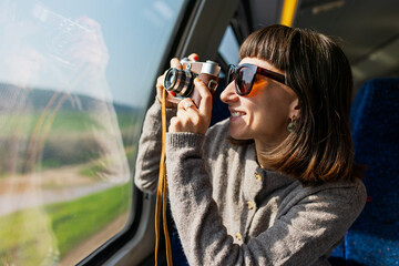 A beautiful, happy young woman takes a photo with a vintage analog camera from a train window. Concept of travel and discovery.