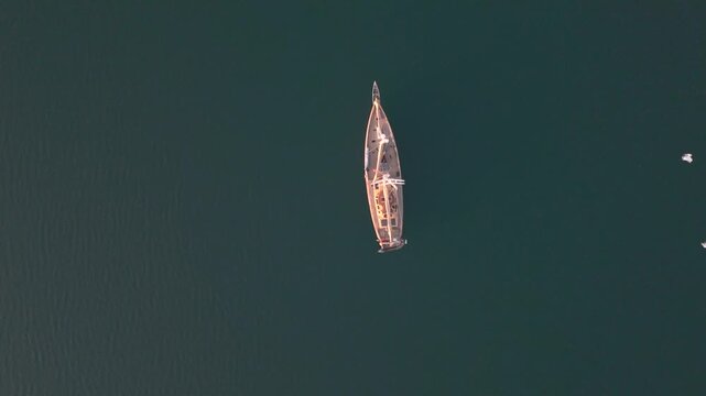 Perpendicular drone shot passes over stationary sailboat floating on green sea