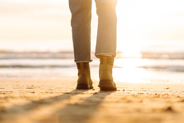 A close-up of a woman's legs in boots against the backdrop of the sea in the sun's rays. Person stands on a sandy beach in boots at sunset. Concepts of hiking, adventure, and walk.