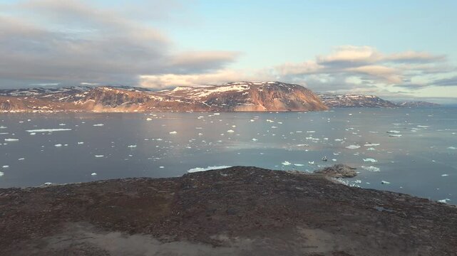 Drone flies over Greenland land toward icy Arctic sea beneath midnight summer light