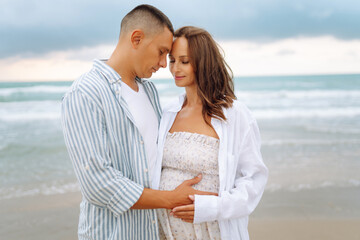 Obraz premium Happy expectant parents stand on the beach and enjoy the seascape. A man and his pregnant wife stroll along the seashore. Concepts of family, expectation, and love.