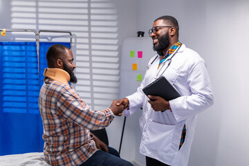 African doctor shakes hands with male patient wearing neck brace in hospital, sharing a moment of comfort and trust.