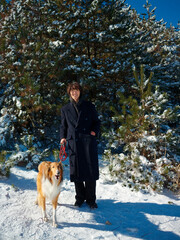Handsome Chinese young man with curly hair in black overcoat smiling with his dog in snow capped mountain forest in sunny winter day, hand in pocket and hold dog leash. Adventure and travel with pet.