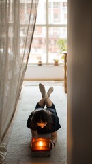 Young woman with dark hair lying on a soft carpet looks into a glowing vintage light box in a cozy room with a large window and natural daylight