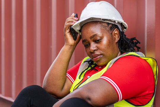 Exhausted female African construction worker in hard hat and safety vest sitting against a red container, looking stressed and tired while holding a phone during a break at a logistics site.