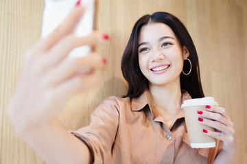 Young, happy, beautiful, stylish Asian woman in brown shirt smiling and taking selfie photo with smartphone, standing in front of wooden wall. Digital technology, social media, modern lifestyle