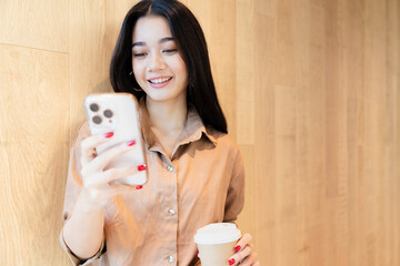 Young, happy, beautiful, stylish Asian woman in brown shirt hold and look at smartphone, smile, coffee cup in the other hand, stand in front of wooden wall. Digital technology, modern social lifestyle