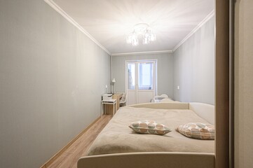 A narrow bedroom with light grey walls, beige bed, patterned pillows, wooden flooring, a desk with a lamp, a chandelier, and a double door window letting in natural light