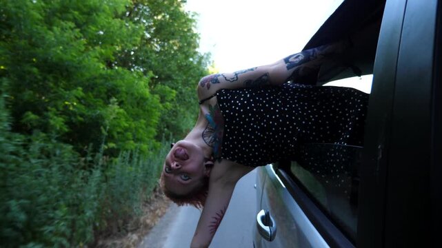Joyful punk girl with tattoos leaning out of auto window and having fun during journey. Happy hippie woman with pink hair enjoying trip while riding on car through country road. Travel concept