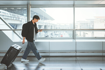 Young Asian man with backpack pulling a suitcase and using a smartphone while walking in a modern...