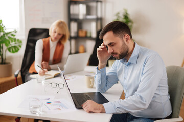 Two coworkers work in an office setting. One person appears stressed and tired while focusing on a laptop. Another person is writing notes while seated nearby.