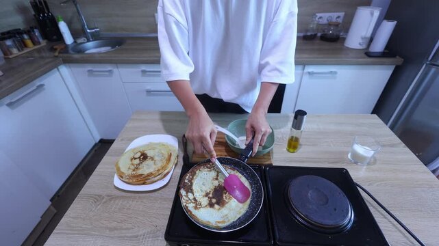A cook gently lifts and flips a golden crepe using a pink spatula, ensuring even browning. Great for mastering delicate flipping techniques in a cozy home kitchen setting.