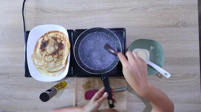 Top-down view of hands brushing oil onto a hot pan, then pouring fresh batter to cook the next pancake. Neat setup with stacked pancakes nearby perfect for batch cooking.