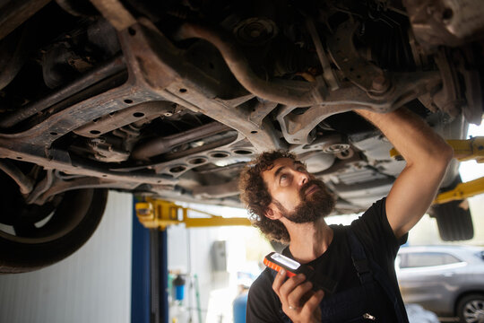 A skilled mechanic examines the undercarriage of a lifted car while holding a diagnostic tool in an auto repair shop during the afternoon. - Powered by Adobe