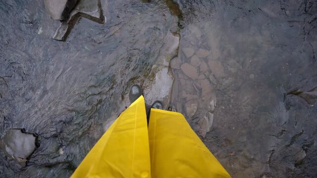 White woman pov stepping on stones, closeup boots and hem of raincoat, slippery rocks and shallow water, careful footing, field scientist checking flow, rugged