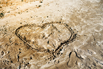 A heart shape drawn in wet sand on a beach, surrounded by natural textures, footprints, and gentle wave patterns. Warm sunlight highlights the sandy surface, creating a romantic  atmosphere.