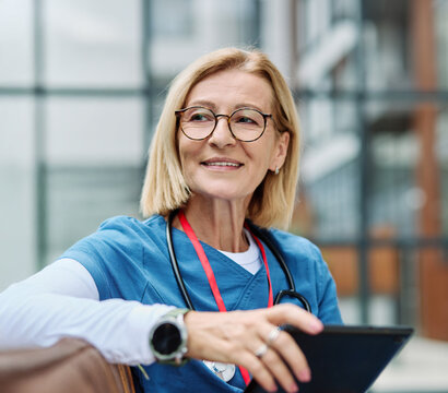 Nurse in scrubs enjoying coffee and using a tablet computer  outside a modern medical facility during a sunny day - Powered by Adobe