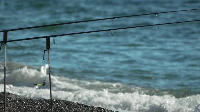 Fishing sea rods waiting for a bite by the deep blue ocean water waves crashing on the rocky beach shore.