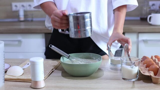 A person hands sifts flour through a metal sieve into a green bowl of wet batter. They then whisk the mixture, combining the dry and wet ingredients to create a smooth, consistent batter for baking.
