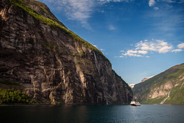 Summer view of Geirangerfjord, Norway. Steep rocky cliffs with green slopes and waterfalls rise above calm blue water, with a passenger ship sailing through the iconic Scandinavian fjord.