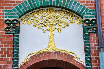 Dekoracyjna złota płaskorzeźba drzewa umieszczona na jasnej ścianie, w otoczeniu czerwonej ceglanej arkady i zielonych kolumn z płytek. © Pol Badger