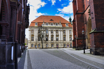 Historyczny rynek z brukowanym placem, budynek ratusza z białą fasadą i ceglanymi łukami po obu stronach – katedra i stary ratusz na rynku w Legnicy. © Pol Badger