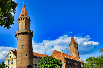 Zabytkowy zamek z cegły z dwiema okrągłymi wieżami pokrytymi czerwoną dachówką, na tle intensywnie błękitnego nieba - Zamek Piastowski w Legnicy © Pol Badger