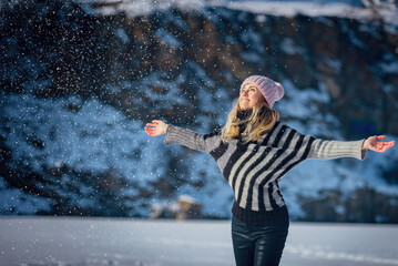 Happy young woman playing with snow on sunny winter day in mountains. Joyful girl playing in the snow, wearing a sweater. Bright mountain landscape and sunny winter atmosphere