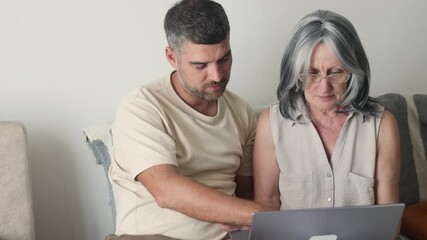 Senior Online Shopping is easy. Son explains Senior Online Shopping to mother. During Senior Online Shopping they choose clothes and use credit card on laptop in living room now.