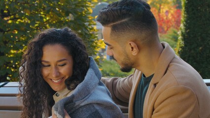 Fototapeta premium Arabian ethnic Hispanic couple in autumn park lovely caring man covering woman with plaid blanket warmth cold weather protection boyfriend cuddling with girlfriend talking smiling sitting on bench