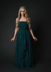 Fototapeta premium Full length portrait of blonde female model wearing royal green ballgown, fantasy crown. Standing, walking pose, front view isolated figure on dark studio background.