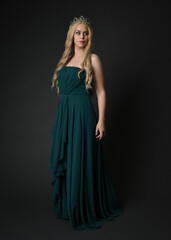 Fototapeta premium Full length portrait of blonde female model wearing royal green ballgown, fantasy crown. Standing, walking pose, front view isolated figure on dark studio background.