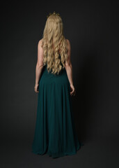 Fototapeta premium Full length portrait of blonde female model wearing royal green ballgown, fantasy crown. Standing, walking away pose, back view isolated figure on dark studio background.