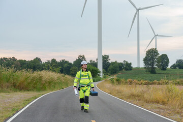 Obraz premium Caucasian adult male engineer walking on rural road carrying toolbox, wind turbines ahead, renewable electricity generation inspection, clean green energy technology, teamwork, outdoor safety uniform