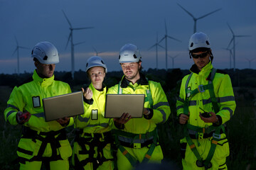 Caucasian adult male female engineer team analyzing laptop data in wind farm for wind turbine maintenance and electricity generation at dusk safety uniform, teamwork for renewable clean energy project