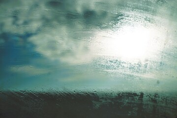 Abstraktes Landschaftsmotiv mit gefrorener Eisbedeckter Glasscheibe vor gr&uuml;nem Feld und blauem Himmel mit blau-wei&szlig;em Wolkengebilde und Licht bei Sonne, Frost und Sturm am Nachmittag im Winter