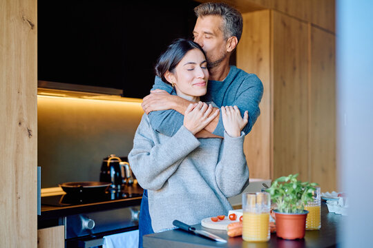 Happy couple sharing an intimate moment in their modern kitchen, the man kissing the woman's forehead during breakfast preparation