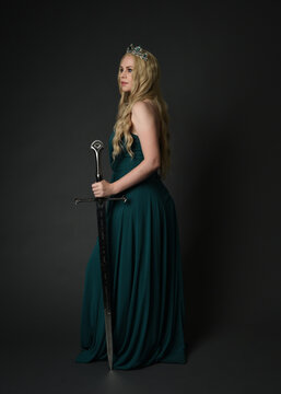 Full length portrait of blonde female model wearing royal green ballgown, fantasy crown. Standing pose walking away from camera, back view, holding sword weapon. isolated figure on dark studio backgro