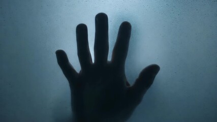 Black silhouette of human hand pressed against frosted glass with cold blue light and minimal background. Concept of isolation and mystery