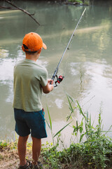 A boy in an orange cap standing alone on the river bank holding a fishing rod and watching the water