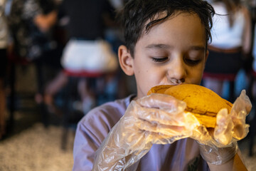 A young boy with short brown hair enjoys a large burger while wearing transparent gloves, seated in a bustling restaurant with people in the background and bright warm lighting