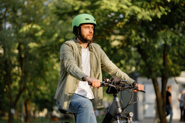 Fototapeta premium Man rides bicycle in city park during sunny day with bright green helmet and casual clothing while trees surround him