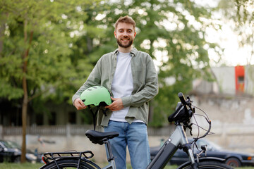 Young man holds a green bicycle helmet while standing next to his bike in a park during daytime