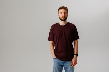 Young man stands against plain background in casual outfit with hands in pockets at studio during daytime