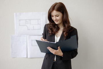 Young woman writes notes while standing near building plans on wall