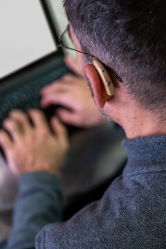 Person with hearing aid and glasses intently typing on a laptop keyboard, focusing on work, digital accessibility, and technology