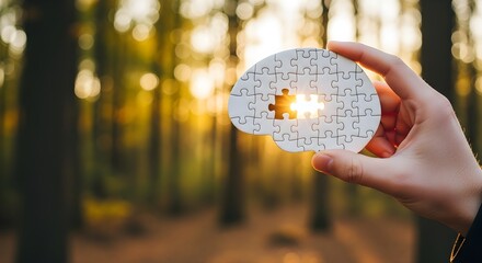 Hand holding a human brain jigsaw puzzle piece against a blurred autumn forest background with bright sunlight shining through the missing gap